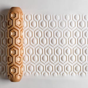 May include: A wooden rolling pin with a geometric honeycomb pattern. The pin is light brown and cylindrical, with a repeating design of interconnected hexagons. The pattern is imprinted on a white surface, creating a textured effect.