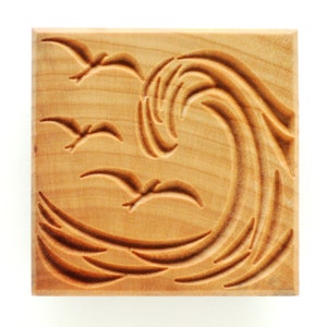 May include: A wooden carving of a wave with three birds flying above it. The carving is on a square piece of wood with a light brown color.