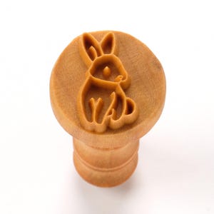 Pode incluir: Um carimbo de madeira castanho claro com um desenho de coelho. O carimbo tem um topo redondo com a gravura do coelho e uma pega cilíndrica. O coelho é retratado sentado com as orelhas erguidas.