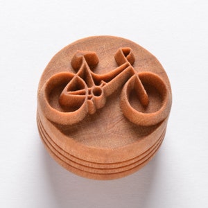 以下が含まれることがあります： 自転車の彫刻が施された木製のスタンプ。スタンプは茶色で丸みを帯びています。