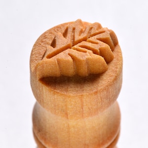May include: A wooden stamp with a carved leaf design. The stamp is light brown and has a smooth, polished finish.