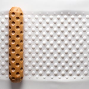 May include: A light brown wooden rolling pin with circular holes lies next to a white surface covered in small, raised dots. The rolling pin is cylindrical in shape.