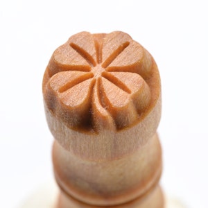 May include: Close-up of a wooden stamp with a floral design. The stamp is light brown and features a detailed, carved flower pattern on its top surface. The handle is cylindrical and also made of wood.