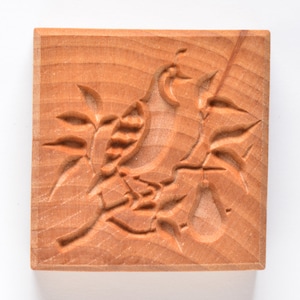 May include: A square, wooden cookie mold with a carved design of a bird perched on a branch with leaves and a pear. The wood has a light brown color and visible grain.