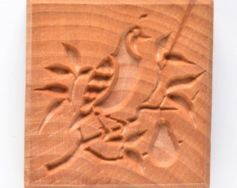May include: A square, wooden cookie mold with a carved design of a bird perched on a branch with leaves and a pear. The wood has a light brown color and visible grain.