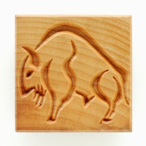 May include: A square, wooden stamp with a carved design of a bison. The bison is depicted in a stylized, artistic manner, with curved lines and detailed features. The wood has a light brown color.