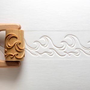 May include: A wooden rolling pin with a wave pattern is shown next to a white surface with the same wave design. The rolling pin has a natural wood finish and is designed to imprint the wave pattern onto dough or clay.