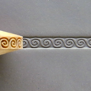 May include: A wooden rolling pin with a carved spiral design is being used to create a border on a gray clay surface.
