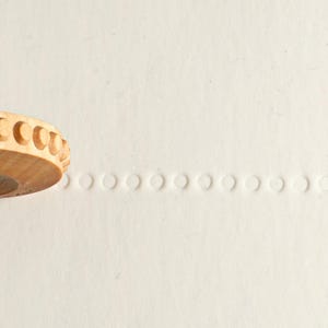 May include: A wooden roller with a row of small, round bumps along the edge. The roller has been used to create a line of small, round indentations on a white surface.