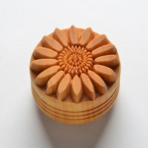 May include: A wooden sunflower-shaped object with detailed carving. The top features a sunflower design with petals and a textured center. The sides have horizontal grooves. The object is light brown in color and sits on a white surface.