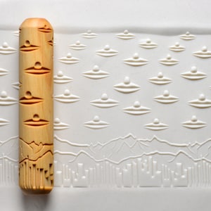 May include: A wooden rolling pin with a carved design of flying saucers and a cityscape. The rolling pin is made of light brown wood and has a smooth, polished finish.