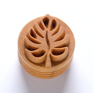May include: A wooden stamp with a carved leaf design. The stamp is brown and has a rounded shape.