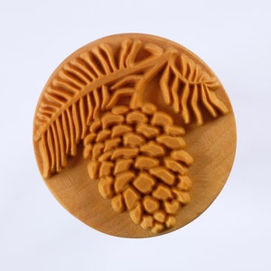 May include: A wooden round ornament with a carved design of a pine cone and a pine branch.