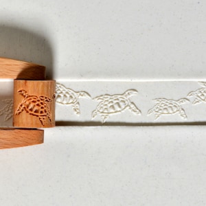 May include: A wooden rolling pin with a turtle design is used to create a pattern of turtles on a white surface.