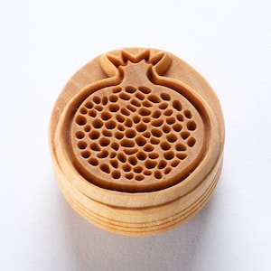 May include: A wooden stamp with a detailed carving of a pomegranate. The stamp is made of light brown wood and has a rounded shape.