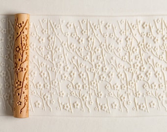 May include: A wooden rolling pin with a floral design is shown next to a textured surface with an embossed floral pattern. The rolling pin is light brown and features a carved floral motif. The embossed pattern is white and features a repeating floral design.