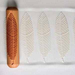 May include: A wooden rolling pin with a leaf design. The rolling pin is used to create leaf patterns in dough or clay. The rolling pin is on a white surface with three leaf impressions.