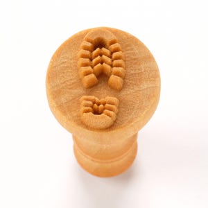 May include: A light brown, wooden stamp with a boot print design. The stamp features a detailed imprint of a boot sole, including the tread pattern. The handle is cylindrical, and the stamp is circular.