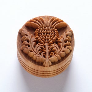 May include: A wooden stamp with a carved design of a thistle flower. The stamp is round and has a brown color.