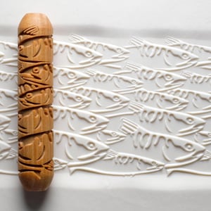 May include: A wooden rolling pin with carved fish designs. The rolling pin is light brown and cylindrical, with a series of fish faces carved around its circumference. The background features a white surface with a repeating pattern of fish.