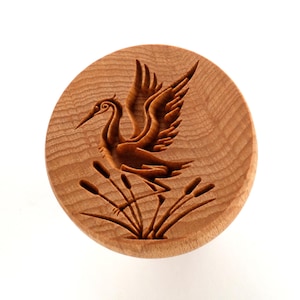 May include: A round, wooden coaster with a detailed carving of a crane in flight above cattails. The coaster is made of light brown wood and has a natural wood grain pattern. The crane is depicted with outstretched wings and a long beak.