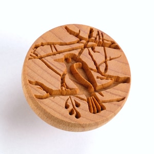 May include: A round wooden cookie cutter featuring a carved design of a bird perched on a branch with berries. The cutter is made from light brown wood and has a smooth, polished finish.