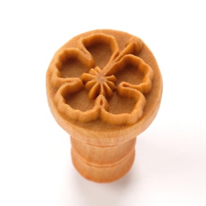 以下が含まれることがあります： 詳細なハイビスカスの花の模様が施された、明るい茶色の木製クッキー型。スタンプは花が付いた丸い上部と円筒形のハンドルが付いています。画像は白い背景にあります。