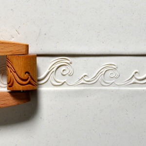 May include: A wooden rolling pin with a carved wave design. The rolling pin is used to create decorative patterns in clay or dough.