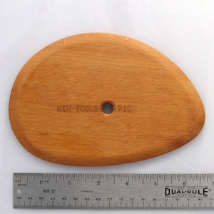 May include: A light brown wooden sculpting tool with a hole in the centre. The tool is oval shaped and has the text "MKM TOOLS W2C" engraved on the surface. The tool is measured against a ruler.