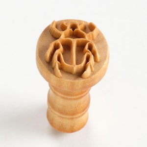 May include: A wooden stamp featuring a raised design of a scarab beetle. The stamp is light brown and has a cylindrical handle.