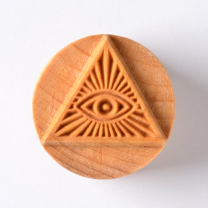 May include: A round, light-brown wooden stamp featuring a carved design of an eye within a triangle, surrounded by radiating lines. The stamp has a natural wood grain texture and is set against a white background.