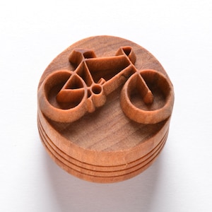 May include: A wooden stamp with a carved design of a bicycle. The stamp is round and has a brown color.