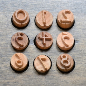 May include: A set of nine circular wooden stamps arranged in a three-by-three grid. Each stamp features a unique raised numeral or symbol carved into the light brown wood surface, displaying characters that resemble various numerical digits.