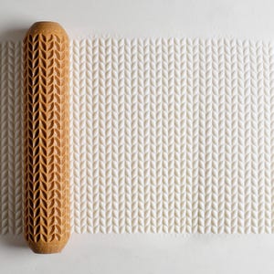 May include: A wooden rolling pin with a carved leaf pattern rests next to a white surface with an embossed leaf pattern. The rolling pin is light brown and cylindrical. The embossed pattern is a repeating design.