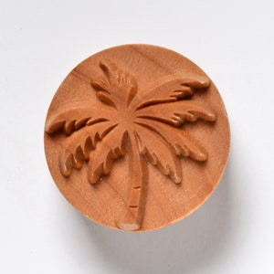 Könnte beinhalten: Ein Holzstempel mit einer detaillierten Schnitzerei eines Palmenbaums. Der Stempel ist rund und hat eine hellbraune Farbe.