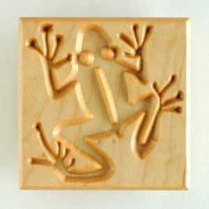 May include: A wooden block with a carved frog design. The frog is facing the right side of the block and has a detailed design with raised lines.