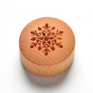 以下が含まれることがあります： 雪の結晶が彫刻された丸い木製のクッキーカッター。カッターは明るい茶色の木でできており、滑らかで磨かれた仕上げになっています。