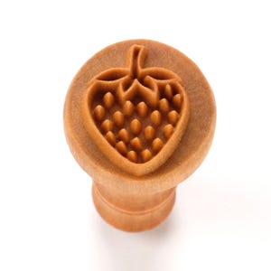 May include: A wooden stamp with a strawberry design. The stamp features a detailed strawberry outline with a textured surface, and a handle for easy use. The stamp is light brown in color and is set against a white background.