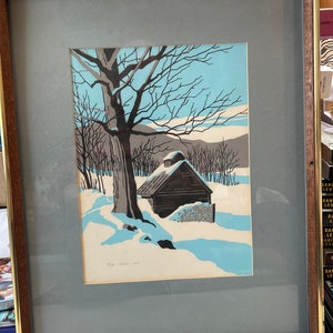 May include: A framed print of a winter scene with a small cabin in the woods. The cabin is brown with a white roof and is surrounded by snow-covered trees. The sky is blue and there are clouds in the distance. The print is signed "B.C. Pastel 1977".