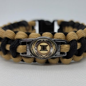 Op de afbeelding: Een zwart en tan paracord armband met een zilveren gesp en een ronde metalen bedel met een gestileerd symbool. De bedel heeft een gouden achtergrond met zwarte en zilveren details.