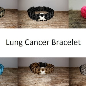 May include: A set of six paracord bracelets with a white ribbon design. The bracelets are in various colors, including black, pink, teal, brown, and gray. The bracelets are displayed on a wooden surface. Lung Cancer Bracelet.