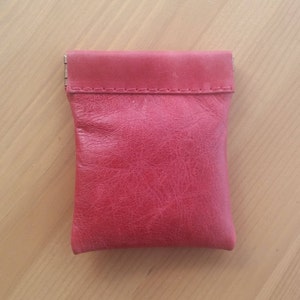 May include: A small, red leather pouch with a flap closure. The pouch is made of textured leather and has a stitched seam around the top edge.