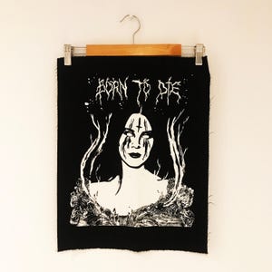 May include: Black fabric patch featuring a white graphic of a woman with dark hair and a cross on her forehead, with the words "BORN TO DIE" in stylized text. The patch is suspended from a wooden hanger.
