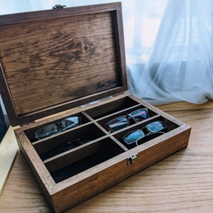 May include: A wooden glasses case with a hinged lid and multiple compartments. The case is stained a dark brown color and contains several pairs of eyeglasses. The text "Love Hals" is visible on the inside of the lid.