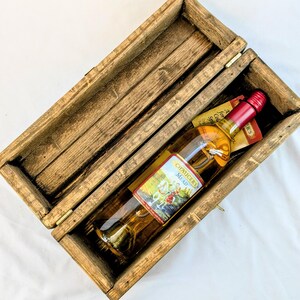 May include: A bottle of Chaucer's Mead with a red cap and label inside a rustic wooden box. The box is open, revealing the bottle and a small tag. The box has a hinged lid and is made of weathered wood.