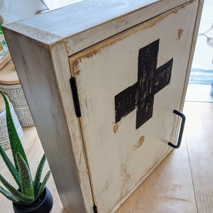 May include: A distressed white wooden cabinet with a black cross on the door. The cabinet has a black handle and hinges. The wood has a worn, rustic appearance, with visible grain and distressed edges. The cabinet is likely for storing medical supplies.