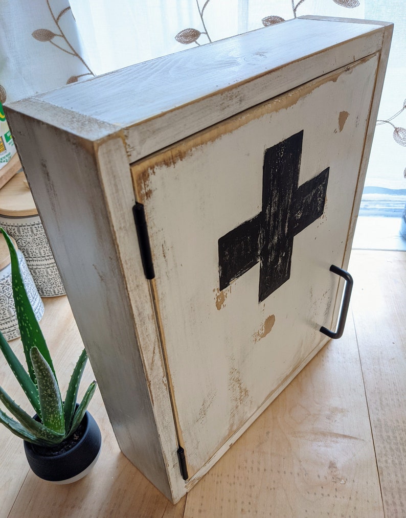May include: A distressed white wooden cabinet with a black cross on the door. The cabinet has a black handle and hinges. The wood has a worn, rustic appearance, with visible grain and distressed edges. The cabinet is likely for storing medical supplies.