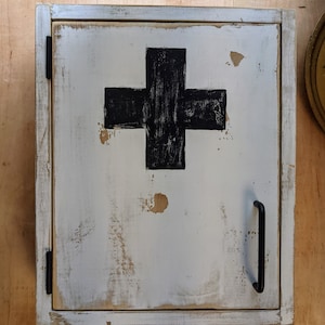 May include: A distressed white wooden cabinet with a black cross on the door. The cabinet has a black handle and hinges. The paint is chipped, giving it a vintage look. The cabinet is likely for storing medical supplies.