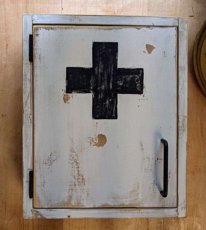 May include: A distressed white wooden cabinet with a black cross on the door. The cabinet has a black handle and hinges. The paint is chipped, giving it a vintage look. The cabinet is likely for storing medical supplies.