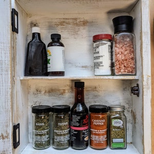 May include: A white, distressed wooden spice rack with two shelves. The top shelf holds two bottles and two jars. The bottom shelf contains four jars of spices and a bottle of soy sauce. The rack is open, revealing the contents.